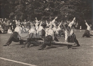 Sepia photo of women performing a side-plank while wearing uniforms consisting of a bib collar, white blouse, and pleated skirt.