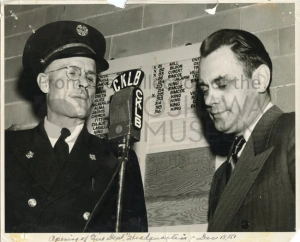 Black and white photo of two Caucasian men, standing beside a microphone. One is wearing a uniform with a hat, and the other is wearing a suit.