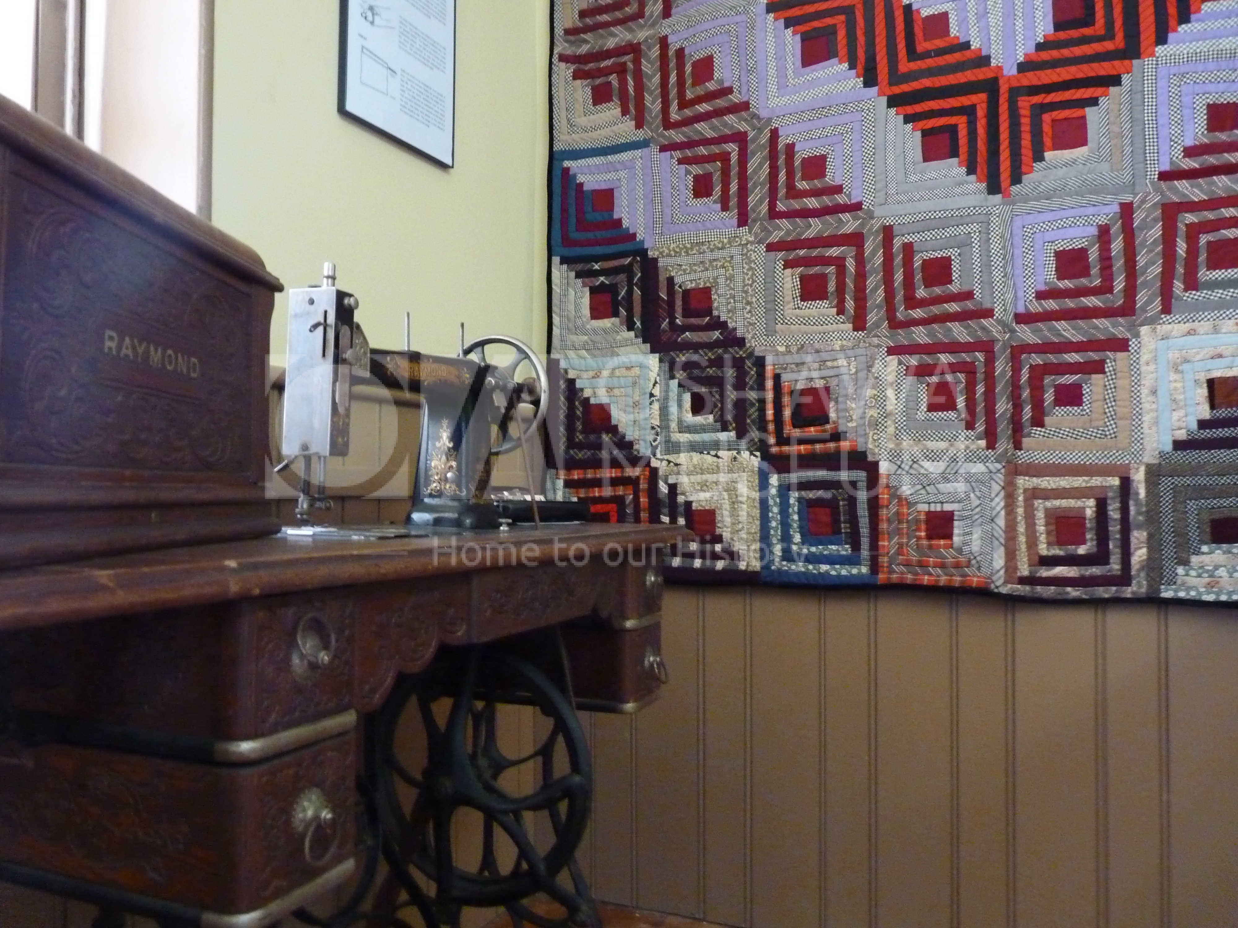 A room with a colourful quilt hanging from the wall and a wooden and metal sewing machine in front of it